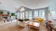 Located at 3543 Idlewood Ter in Indianapolis, IN, this single-floor living space offers a thoughtfully designed open-concept layout that maximizes both functionality and visual appeal. The room is anchored by soaring vaulted ceilings with a prominent arched window that floods the interior with natural light, complemented by a ceiling fan and recessed lighting throughout. Light gray walls and wood-look flooring create a clean, cohesive aesthetic across the entire space. The living area features comfortable white upholstered seating accented with colorful throw pillows, arranged around a classic wooden coffee table atop a plush area rug. A wall-mounted flat-screen TV and wooden media console define a dedicated media zone, with French doors providing access to an adjacent area. A stone fireplace with a wooden mantel serves as a striking architectural focal point, adding warmth and character. A defined dining area with a glass-top table and upholstered chairs sits nearby. Large multi-pane windows line the walls, offering views of the outdoor pool and surrounding greenery. Decorative plants and tasteful wall art are placed throughout, enhancing the bright, welcoming ambiance of this well-appointed space.