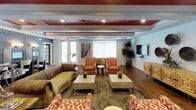 This single-floor community amenity space combines functional design with bold aesthetic choices, creating an inviting environment for gathering and recreation. Rich, warm hardwood flooring flows continuously throughout, unifying each area with a cohesive, upscale feel.

The kitchen is well-appointed with dark espresso cabinetry, a subway tile backsplash, black appliances including a range and oven, a stainless steel side-by-side refrigerator with ice and water dispenser, and ample counter space. A large window fitted with white horizontal blinds floods the area with natural light, while a dedicated coffee station adds a welcoming touch.

The lounge area is a standout, featuring a dramatic tray ceiling accented with bold red insets and recessed lighting that creates a lively, energetic atmosphere. Eclectic furnishings — including patterned armchairs, neutral sofas, and decorative side tables — are arranged across a textured area rug, offering multiple seating configurations. Wall-mounted flat-screen televisions, curated artwork, and ornamental wall accents contribute to the space's distinctive character. Large windows provide generous natural light and a connection to the outdoors.

An open transitional area with structural columns leads to additional rooms, including what appears to be a fitness center and a vanity or grooming station, making this a comprehensive and versatile community hub.