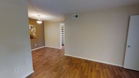 This single-floor residence offers a clean and functional layout designed for comfortable everyday living. The main living and dining area is finished with warm-toned hardwood-style flooring that runs throughout, adding a sense of cohesion and warmth to the space. Neutral beige and tan wall tones create a versatile backdrop suitable for a variety of furnishing styles. A textured ceiling adds subtle depth overhead, while a pendant light fixture provides focused illumination over the dining area. Ventilation is well-considered, with visible HVAC vents integrated into both the wall and ceiling. The open layout allows for a natural flow between the living and dining spaces, with a doorway leading further into the residence — offering a glimpse of additional rooms beyond. White trim and baseboards provide crisp contrast against the warmer wall tones, contributing to a tidy, well-maintained aesthetic. Decorative wall art accents one corner of the living area, hinting at the personalization the space readily accommodates. Overall, the residence presents a straightforward, move-in-ready environment with practical finishes and a welcoming atmosphere throughout its single level.