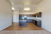 Located at 1275 Mike St in Dickinson, North Dakota, this one-bedroom, one-bathroom unit offers a well-designed single-level layout with a comfortable and functional floor plan. The open-concept kitchen and living area serves as the heart of the home, highlighted by soaring vaulted ceilings that lend an impressive sense of height and spaciousness. The kitchen is well-appointed with rich, dark wood cabinetry, granite-style countertops, a central island for additional prep space, and a full suite of black appliances including a refrigerator, range, microwave, and dishwasher. Hardwood-style flooring runs throughout the kitchen area, transitioning to plush carpeting in the living room. The living room features a large window that fills the space with natural light and offers views of the surrounding Dickinson landscape, along with a wall-mounted HVAC unit and baseboard heating. The bedroom is generously sized with soft carpeting, a closet, and a sliding glass door that opens to a private balcony with open views. A dedicated laundry area houses a full-size top-loading washer and includes wire shelving above for added storage. The bathroom completes this thoughtfully arranged unit, making it a practical and comfortable residence.