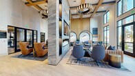 Located at 6851 N 93rd Ave in Glendale, Arizona, this upscale communal living room is a masterclass in contemporary interior design. The space commands attention with its dramatic double-height ceilings, accented by a bold coffered framework finished in warm gold tones and adorned with striking circular and layered pendant light fixtures that create a sophisticated ambiance throughout.

At the heart of the room stands a floor-to-ceiling marble feature wall housing a large flat-screen television above a modern linear fireplace flanked by decorative candle displays — serving as a stunning focal point. Vertical wood slat paneling adds warmth and texture to the surrounding walls, complementing the light-toned wide-plank flooring that runs throughout.

Seating is thoughtfully arranged into distinct zones: rich cognac leather wingback chairs, blue upholstered swivel chairs, and plush sofas are grouped around coffee tables, offering both intimate and social settings. Arched booth-style seating alcoves with integrated greenery line one wall, providing semi-private lounging areas. A separate dining zone features sleek round bistro tables.

Floor-to-ceiling black-framed windows and glass doors flood the space with natural light while offering views of the surrounding Glendale community. The overall aesthetic balances luxury and comfort, making this an inviting and visually dynamic gathering space.
