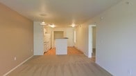 This two-floor space features an open-concept layout on the main level, where the living area and kitchen flow seamlessly into one another. The living area is finished with neutral-toned carpet, creating a warm and comfortable atmosphere, while the kitchen and adjoining spaces transition to hardwood-style flooring. The kitchen is equipped with white appliances, including a refrigerator and range, and is complemented by white cabinetry and a central island that serves as both a workspace and a natural divider between the kitchen and living areas. A pendant light fixture hangs above the island, providing focused task lighting, while additional ceiling-mounted fixtures illuminate the surrounding space. The walls throughout are painted in a clean, neutral white, contributing to a bright and airy feel. A doorway visible from the main living area leads to additional rooms, suggesting a functional and well-organized floor plan across both levels. The overall design is straightforward and practical, with a neutral color palette that offers flexibility for a variety of furnishing styles.