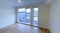 This single-floor space offers a clean, contemporary design defined by an abundance of natural light and an open, airy feel. The main living area is anchored by a striking set of large, floor-to-near-ceiling windows that span much of one wall, framing panoramic views of the surrounding urban landscape and tree-lined scenery beyond. The elevated vantage point enhances the sense of openness, allowing natural light to flood the interior throughout the day.

The flooring throughout features light-toned hardwood-style planks with subtle grain variation, lending warmth and continuity to the space. The walls are finished in a crisp, neutral white, creating a versatile backdrop well-suited to a variety of interior styles. Recessed ceiling lighting provides a polished, unobtrusive overhead illumination to complement the natural light.

The overall aesthetic is minimalist and modern, with smooth wall transitions and clean architectural lines throughout. The combination of expansive glazing, elevated positioning, and neutral finishes creates a bright, welcoming atmosphere with a strong connection to the outdoors.