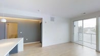 This single-floor space showcases a clean, contemporary design aesthetic defined by a neutral color palette and modern finishes. The main living area features light-toned hardwood flooring that runs throughout, contributing to a bright and open feel. White walls and a smooth white ceiling create a crisp, airy atmosphere, while a ceiling-mounted HVAC vent ensures climate comfort.

A distinctive architectural detail is the recessed alcove or adjoining area, which features contrasting gray walls and warm amber LED cove lighting along the upper edge, adding depth and a modern ambiance to the space. A dark-framed doorway within this zone leads to an additional room or hallway, enhancing the sense of flow between areas.

Natural light is introduced through a window fitted with horizontal blinds on the right side of the space, allowing for adjustable brightness. The walls include multiple electrical outlets positioned at standard heights, offering practical functionality. Overall, the space strikes a balance between minimalist design and warm accent elements, making it a versatile and inviting environment.