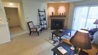 This single-floor space is designed for comfortable, everyday living, with a warm and inviting ambiance throughout. The living area serves as the centerpiece of the home, anchored by a tile-surround fireplace with a classic wood mantel and a rich terracotta accent wall that adds a pop of warmth and visual depth to the room. Track lighting overhead provides adjustable illumination, complementing the natural light that filters in through vertical blinds on a large window or sliding door. Soft wall-to-wall carpeting underfoot contributes to the cozy, relaxed atmosphere of the space. The living area is well-proportioned, offering ample room for seating arrangements and entertaining, with space for accent furniture such as a ladder-style bookshelf for display and storage. The layout is straightforward and practical, making it easy to arrange and personalize. Overall, the space strikes a balance between functional design and homey comfort, with thoughtful details like the decorative mantel display and coordinated furnishings that give it a lived-in, welcoming character.