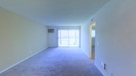 This single-floor unit offers a straightforward and functional layout designed for comfortable everyday living. The main living area is generously proportioned, featuring plush wall-to-wall carpeting that adds warmth underfoot. A large sliding glass door dominates one wall, fitted with vertical blinds that allow for adjustable natural light and providing direct access to an outdoor area. The abundance of natural light from this opening gives the living space an open, airy feel. A wall-mounted air conditioning unit ensures climate control throughout the warmer months, while a floor vent provides additional temperature regulation. The walls are finished in a clean, neutral tone, offering a versatile backdrop for a variety of interior styles. An open doorway visible from the main living area leads to an adjoining room, suggesting a connected and accessible floor plan. The textured ceiling and crisp white trim and baseboards throughout contribute to the clean, well-maintained character of the space. Overall, the unit presents a practical and comfortable living environment with key amenities thoughtfully in place.