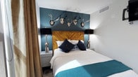 This guest suite is a thoughtfully designed space that blends rustic charm with modern sophistication. The centerpiece of the room is a striking deep teal accent wall that serves as a dramatic backdrop for a collection of mounted antler sculptures, adding a distinctive lodge-inspired aesthetic. A beautifully crafted herringbone-patterned wood headboard anchors the room, lending warmth and texture to the overall design. The bed is dressed in crisp white linens complemented by rich navy throw pillows and a bold teal accent blanket, tying the color palette together cohesively. Flanking the bed are matching nightstands, each topped with elegant chrome-based lamps featuring black shades that provide warm, ambient lighting. The contrast between the cool teal tones and the warm wood elements creates a balanced and inviting atmosphere. Soft drapery along the walls adds a layer of texture and comfort to the room. The space is efficiently laid out to maximize comfort while maintaining a clean, uncluttered feel. Overall, the guest suite delivers a memorable and immersive experience, combining bold design choices with thoughtful furnishings to create a welcoming and visually engaging environment.