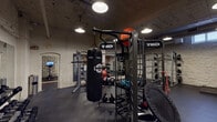 This single-level fitness facility combines an industrial aesthetic with a comprehensive array of professional-grade equipment, creating a functional and immersive workout environment. The exposed whitewashed brick walls, visible ductwork, and concrete ceilings give the space a raw, urban character, while pendant lighting throughout provides focused illumination across each training zone.

The strength training area features a power rack with an extensive plate collection, rubber-matted flooring, and a warm wood-paneled accent wall, alongside a large mirror that enhances the sense of depth. A dedicated functional fitness zone is anchored by a TKO multi-functional training rig, complete with a hanging heavy bag, kettlebells, medicine balls, and resistance cables — catering to a wide range of dynamic training modalities.

The cardio zone is well-appointed with treadmills, elliptical machines, stationary bikes, and a rowing machine, all arranged along a vibrant scenic mural of an open road at sunset, accompanied by wall-mounted screens to keep users engaged during sessions. A separate free weights area offers a full dumbbell rack and stability balls, supported by additional display screens. Throughout the space, rubber flooring ensures durability and safety, making this facility a thorough and thoughtfully organized training environment.