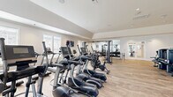 Located at 1090 Summersweet Dr in New Bern, NC, this single-level fitness center is a comprehensively outfitted facility designed to support a full range of workout routines. The main exercise floor is expansive, bathed in natural light from multiple large windows with white blinds, and finished with warm, light-toned hardwood-style flooring throughout. A wall of floor-to-ceiling mirrors along one side visually amplifies the space and provides functional feedback during workouts.

The cardio zone is well-stocked with a row of treadmills, ellipticals, and stationary bikes — several equipped with individual display screens — lined neatly along the windowed walls. The strength training area features a tiered dumbbell rack with a wide range of weights, cable and multi-use machines, a power rack with TRX suspension straps, a heavy bag, medicine balls, and benches to support varied resistance training.

A separate, enclosed room offers a quieter space for stretching, yoga, or low-impact exercise, featuring its own large interior window, wall-mounted shelving for accessories, and a step platform. Crown molding and tray ceiling details add a refined architectural touch throughout, and recessed lighting keeps the environment bright and welcoming. Double glass doors provide easy access to the exterior, enhancing airflow and connectivity to the surrounding community.