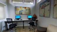 This single-floor business center offers a thoughtfully designed workspace that balances functionality with a refined aesthetic. The room features soaring ceilings with clerestory windows positioned near the top of the walls, allowing natural light to pour in and creating an open, airy atmosphere that makes the space feel expansive and inviting.

The workstation area is equipped with multiple desktop computer setups arranged along a sleek, minimalist white desk, each accompanied by ergonomic mesh office chairs that prioritize comfort during extended use. The light wood-toned flooring adds warmth to the otherwise clean and neutral color palette of the white walls.

Curated artwork elevates the professional environment, including a large abstract canvas in warm earth tones — golds, blues, and oranges — displayed prominently above the workstations, and a vibrant, whimsical painting on the adjacent wall that adds a touch of creative energy to the space. Decorative ceramic pieces further contribute to the polished yet approachable character of the room.

Overall, this business center strikes a balance between a productive work environment and an aesthetically engaging space, making it well-suited for focused individual work or shared use within a professional or community setting.