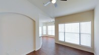 This single-story space makes a striking impression through its vaulted ceilings, which lend a sense of grandeur and openness to the interior. The living area is finished with rich, dark hardwood flooring that runs throughout, providing a warm and cohesive aesthetic against the neutral, cream-toned walls.

A graceful arched doorway serves as a defining architectural detail, elegantly framing the transition between areas and adding a classic design touch. The main living space is well-lit by multiple large windows dressed with horizontal blinds, allowing ample natural light to fill the room while offering privacy control.

Overhead, a ceiling fan is centrally mounted, providing both practical air circulation and a subtle decorative element suited to the vaulted ceiling height. Further into the space, a track lighting fixture highlights an additional living or dining area, which benefits from a bay-style window arrangement that enhances the sense of openness and draws in additional light.

White baseboards and trim provide clean contrast throughout, complementing the overall bright and welcoming ambiance. The open layout and architectural details combine to create a spacious, comfortable environment well-suited for everyday living.