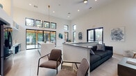 This single-floor space showcases a striking contemporary design centered around an expansive open-concept layout with dramatically high ceilings that amplify the sense of volume throughout. Oversized black-framed windows, including clerestory windows positioned near the ceiling, flood the interior with abundant natural light while framing views of the surrounding outdoor areas and mature trees.

The kitchen is a standout feature, incorporating two distinct islands. The first displays a bold teal geometric-patterned base topped with a white quartz surface and is flanked by warm cognac-colored barstools beneath a brass-and-glass chandelier. The adjacent cabinetry features a sophisticated blue-grey palette complemented by a textured blue zellige tile backsplash, open shelving, and full-size stainless steel appliances. The second island presents a dramatic waterfall marble-veined stone surface with dark cabinetry below.

The lounge area offers a comfortable seating arrangement with a dark sofa, designer accent chair, and built-in walnut and dark cabinetry with open shelving. A dining area with a round wood table and modern chairs provides a transitional gathering space. Large sliding glass doors open directly to an outdoor covered patio, seamlessly connecting the interior to the exterior. Light-toned large-format tile flooring unifies the entire space.