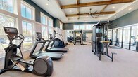 Located at 9500 Grandtree Ave in Venice, Florida, this fitness center offers a generously sized, single-floor layout designed to support a comprehensive workout experience. The space is divided into two distinct zones, seamlessly connected through a glass-panel partition wall that adds a modern, open feel while providing visual separation between areas.

The main workout area is defined by its striking architectural design, featuring elevated tray ceilings accented with warm, exposed wood beams and an oversized ceiling fan. Recessed lighting throughout ensures the space is well-lit at all times. The walls are finished in a bold deep teal above white wainscoting, creating a clean and polished aesthetic. Large windows line the perimeter, flooding the floor with abundant natural light.

The cardio zone is equipped with a row of treadmills, elliptical machines, a stair climber, and stationary spin bikes. The free weights and strength training section features a full power rack with weight plates, a dumbbell rack, and a multi-function cable machine. A dedicated spin and stretching room offers floor-to-ceiling windows framing tranquil water and natural landscape views, creating a serene backdrop for cycling and low-impact training. Light-toned carpet flooring runs throughout, adding comfort underfoot across the entire facility.