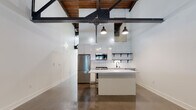 This single-floor space showcases a striking industrial-modern aesthetic, characterized by soaring ceilings that expose original wooden beams and bold black steel trusses overhead. Polished concrete floors run throughout, lending a sleek, cohesive look that complements the unit's raw architectural character.

The open-plan kitchen is a standout feature, outfitted with white cabinetry, full-height subway tile backsplash, open black metal shelving, and stainless steel appliances including a gas range, built-in microwave, refrigerator, and dishwasher. A generous white waterfall island anchors the space, offering ample workspace and serving as a natural divider between the kitchen and the expansive living area. Industrial-style pendant lights hang from the steel trusses above, providing focused illumination.

The main living area is open and versatile, benefiting from polished concrete floors and tall white walls. A separate room, accessible from the main space, features a large black-framed window overlooking an industrial interior, a wall-mounted mini-split HVAC unit, and the same concrete flooring, making it suitable as a bedroom or private office.

The bathroom includes a wood-toned vanity with a white countertop, a mirror, and polished concrete flooring consistent with the rest of the unit. The overall ambiance is clean, modern, and industrial throughout.