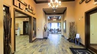 This single-story space presents an inviting lobby and corridor that combines warm aesthetics with functional design. The entryway is finished with natural slate-look tile flooring in earthy tones of brown, gray, and rust, lending a grounded, sophisticated feel underfoot. Coffered ceilings with dark wood trim detailing add architectural depth and visual interest overhead, while an elegant chandelier serves as a focal point, casting warm ambient light throughout the space.

The walls are painted in soft, warm beige tones that complement the rich wood accents found on trim work and sliding glass doors to the left, which appear to lead to an office or administrative area. Decorative elements include framed artwork, a bamboo plant arrangement in a floor planter, and a small console table, all contributing to a polished yet welcoming atmosphere.

The corridor leads naturally toward a lounge area at the far end, where comfortable seating, a coffee table, and large windows with draping curtains allow natural light to filter in. The overall ambiance of the space is professional yet warm, suggesting a well-maintained communal or commercial reception area designed to make visitors feel at ease upon arrival.