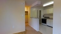 This single-floor residence offers a practical and well-organized layout designed for comfortable everyday living. The interior is finished with neutral, light-toned walls throughout, creating a bright and cohesive atmosphere that flows naturally from room to room.

The kitchen is positioned along one side of the space and is defined by a half-wall pass-through that opens to the main living area. White upper cabinetry provides generous storage, and the kitchen is equipped with a gas range. Tile flooring runs through the kitchen area, offering durability and ease of maintenance. A fluorescent overhead light ensures the workspace is well-lit.

Transitioning into the living area, soft carpet flooring adds warmth underfoot. A structural beam detail is visible overhead, adding a subtle architectural element to the ceiling. The half-wall pass-through enhances the sense of openness between the kitchen and living spaces, allowing natural sightlines across the unit.

A dedicated walk-in closet is accessible from the hallway, offering notable built-in shelving and hanging rod storage. A bifold-door closet is also visible nearby, further contributing to the unit's overall storage capacity. The layout is efficient and well-suited for everyday functionality.