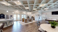Located at 100 Pinckney Marsh Ln in Port Royal, SC, this single-story community clubhouse offers a thoughtfully designed gathering space with a range of amenities to serve its residents. The centerpiece of the facility is a spacious main lounge area distinguished by an elegant coffered ceiling with recessed lighting, rich hardwood-style flooring, and an abundance of natural light streaming through multiple sets of glass French doors. Comfortable seating arrangements invite relaxed socializing, while the open layout creates a welcoming, airy atmosphere throughout.

Adjacent to the lounge is a generously sized game room, equally impressive with its matching coffered ceiling and large windows dressed with soft drapery. A full-size pool table anchors the space, complemented by a wall-mounted television and a built-in window bench — ideal for casual recreation and entertainment.

A dedicated business center provides two workstations with computers, built-in cabinetry, and a shared printer, offering a quiet and functional space for residents' professional needs. The communal kitchen features stainless steel appliances, white cabinetry, and direct access to an outdoor patio area. A well-maintained laundry facility rounds out the amenities, equipped with multiple washers and dryers, a wall-mounted television, and a checkered tile floor. Connecting corridors with glass-panel doors maintain visual openness throughout the entire facility.