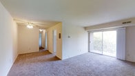 This single-floor space features an open living area with wall-to-wall carpet flooring in neutral tones, creating a clean and understated foundation throughout. A sliding glass door along one wall provides direct access to a private balcony, where views of surrounding greenery are visible through the glass, allowing natural light to filter into the main living space. The exterior brick detail framing the balcony adds a classic architectural touch.

The layout connects the main living area to an interior hallway, which leads to additional rooms beyond. A ceiling fan is visible in an adjacent area, indicating a secondary living or dining zone. The hallway features multiple doorways, suggesting access to bedrooms or additional functional spaces, along with what appears to be a utility panel or thermostat mounted on the wall.

The overall color palette is light and neutral, with cream-colored walls and standard white trim throughout. The space maintains a straightforward, practical design with consistent flooring and a well-connected floor plan that allows for easy movement between areas. The single-floor layout ensures all spaces are conveniently accessible.