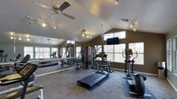 This fitness center is thoughtfully designed to support a full range of workout routines in a bright, open environment. The space features vaulted ceilings that lend an airy, spacious feel, complemented by a ceiling fan that promotes air circulation during workouts. Track lighting runs along the ceiling, ensuring the room is well-lit throughout. Large windows line the walls, allowing ample natural light to fill the space and providing views of the surrounding outdoor area. A full-length mirrored wall along one side visually expands the room while allowing users to monitor their form during exercise. The fitness center is furnished with a variety of cardio equipment, including treadmills and elliptical machines, as well as a multi-station strength training unit for resistance-based workouts. A wall-mounted flat-screen television adds an entertainment element to the space. The flooring consists of durable, textured carpet suited for fitness use. The overall ambiance is clean, functional, and welcoming, making it a practical and comfortable facility for daily exercise.