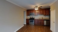 This single-floor residence offers a thoughtfully designed layout that combines functionality with classic style. The kitchen serves as a central highlight, showcasing rich cherry wood cabinetry that extends to the upper walls, providing generous storage space. Granite-style countertops complement the warm tones of the cabinetry, while a full suite of stainless steel appliances — including a gas range, over-the-range microwave, dishwasher, and sink — equips the space for everyday cooking needs. The kitchen flows into an open adjoining area, creating a connected and airy feel throughout. Warm hardwood-style flooring runs seamlessly across the space, adding depth and a sense of continuity. Crown molding along the ceiling lends a refined, finished touch to the interior. A ceiling fan with an integrated light fixture provides both comfort and illumination. Neutral-toned walls keep the space feeling bright and versatile, offering a clean backdrop that suits a variety of interior styles. The overall ambiance is practical yet inviting, with classic design elements that make this a comfortable and well-equipped living space.