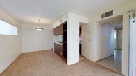 This single-floor residential unit offers a practical and functional layout that maximizes the use of its available space. The open-concept design seamlessly connects the living area to the kitchen, creating a cohesive and inviting environment. The living area is finished with wall-to-wall carpet in a neutral tone, lending warmth and comfort underfoot, while the kitchen transition area features tile flooring for easy maintenance. The kitchen is equipped with dark wood cabinetry featuring open shelving, providing ample storage and display space. A built-in cooktop is integrated into the cabinetry unit, which serves as a natural divider between the living and kitchen spaces. A pendant light fixture hangs above the living area, adding a subtle decorative touch to the otherwise clean, understated aesthetic. The textured ceiling and HVAC vents throughout speak to a well-considered climate control system. White walls and neutral finishes keep the space feeling bright and open. A paneled door leads to an additional room, suggesting further private living quarters beyond the main area. Overall, the unit presents a straightforward, well-maintained living environment suited to a range of occupants.