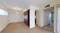 This single-floor residential unit offers a practical and functional layout that maximizes the use of its available space. The open-concept design seamlessly connects the living area to the kitchen, creating a cohesive and inviting environment. The living area is finished with wall-to-wall carpet in a neutral tone, lending warmth and comfort underfoot, while the kitchen transition area features tile flooring for easy maintenance. The kitchen is equipped with dark wood cabinetry featuring open shelving, providing ample storage and display space. A built-in cooktop is integrated into the cabinetry unit, which serves as a natural divider between the living and kitchen spaces. A pendant light fixture hangs above the living area, adding a subtle decorative touch to the otherwise clean, understated aesthetic. The textured ceiling and HVAC vents throughout speak to a well-considered climate control system. White walls and neutral finishes keep the space feeling bright and open. A paneled door leads to an additional room, suggesting further private living quarters beyond the main area. Overall, the unit presents a straightforward, well-maintained living environment suited to a range of occupants.