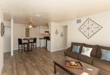 Located at 900 Country Club Dr SE in Rio Rancho, New Mexico, this single-floor unit offers a well-organized and functional layout throughout. The open-concept living area features wood-style plank flooring and a textured ceiling, creating a casual yet comfortable atmosphere. A half-wall partition separates the living space from the kitchen, which is equipped with white cabinetry, black appliances including a refrigerator and range, and ample counter space. A dedicated dining nook sits adjacent to the kitchen, accented by a ceiling light fixture and enough room for a bistro-style table and chairs. The kitchen extends into a utility area housing a stacked washer and dryer, adding in-unit laundry convenience. The bathroom features a white vanity with storage cabinetry, a well-lit mirror, and a combined tub and toilet area. The two bedrooms are carpeted and filled with natural light from windows dressed with sheer curtains. The primary bedroom includes a door leading to an exterior access point, while the secondary bedroom is versatile enough to serve as a guest room or home office. Throughout the unit, neutral tones and clean finishes create a cohesive and move-in-ready living environment.