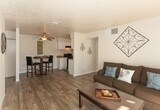 Located at 900 Country Club Dr SE in Rio Rancho, New Mexico, this single-floor unit offers a well-organized and functional layout throughout. The open-concept living area features wood-style plank flooring and a textured ceiling, creating a casual yet comfortable atmosphere. A half-wall partition separates the living space from the kitchen, which is equipped with white cabinetry, black appliances including a refrigerator and range, and ample counter space. A dedicated dining nook sits adjacent to the kitchen, accented by a ceiling light fixture and enough room for a bistro-style table and chairs. The kitchen extends into a utility area housing a stacked washer and dryer, adding in-unit laundry convenience. The bathroom features a white vanity with storage cabinetry, a well-lit mirror, and a combined tub and toilet area. The two bedrooms are carpeted and filled with natural light from windows dressed with sheer curtains. The primary bedroom includes a door leading to an exterior access point, while the secondary bedroom is versatile enough to serve as a guest room or home office. Throughout the unit, neutral tones and clean finishes create a cohesive and move-in-ready living environment.
