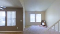 This two-story residence offers a welcoming and functional layout designed for comfortable everyday living. The main living area is generously proportioned, featuring plush wall-to-wall carpeting that creates a warm and inviting atmosphere. Multiple large windows dressed with vertical blinds allow abundant natural light to fill the space, contributing to an airy, open feel. The walls are finished in neutral tones, with a warm taupe accent wall adding depth and visual interest to the living area. A transition zone connects the carpeted living space to an adjacent area with hard-surface wood-look flooring, providing a practical and stylish delineation between spaces. The textured ceiling adds a subtle architectural detail throughout. The two-floor layout provides a natural separation between living and private areas, offering both functionality and privacy. Overall, the space maintains a clean, neutral palette that serves as a versatile canvas, making it well-suited for a variety of furnishing styles and personal aesthetics. The abundance of natural light and open sightlines give the home an expansive, comfortable character.