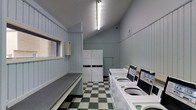 This shared laundry facility is a practical, single-floor space designed to serve the needs of multiple users efficiently. The room is lined with light blue vertical wood paneling that lends a clean, classic aesthetic to the interior. A bold green-and-white checkered vinyl floor anchors the space with a timeless, easy-to-maintain finish.

Along one wall, a row of modern top-load washing machines with digital control panels provides ample laundry capacity. Adjacent to these, two stacked washer-dryer units offer additional flexibility for drying needs. A wide, flat folding counter runs along the opposite wall, providing a generous surface for sorting and folding laundry.

A small window allows a degree of natural light to filter into the room, complemented by bright fluorescent overhead lighting that ensures the space is well-illuminated at all times. A wall-mounted utility box and a security camera add functional and safety-oriented features to the room. The vaulted ceiling gives the space an unexpectedly open feel, preventing the room from feeling cramped despite its focused purpose. Overall, this laundry facility is thoughtfully arranged to maximize usability, comfort, and convenience for its users.