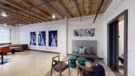 This single-floor loft unit showcases a compelling fusion of industrial heritage and contemporary design. Throughout the space, exposed wood-beam ceilings with visible ductwork and piping preserve an authentic raw aesthetic, while polished terrazzo floors provide a refined, durable foundation underfoot. Whitewashed brick and concrete columns punctuate the open layout, adding structural character.

The expansive living area features a tufted leather sofa, marble coffee table, and French doors opening to a private balcony with outdoor views. A gallery-like wall displays large-scale vivid artwork, with a built-in bench seating area accented by teal accent tables. The modern kitchen is outfitted with stainless steel appliances, white cabinetry, subway tile backsplash, and a striking live-edge wood dining table with leather bar stools.

Two bedrooms offer a calm, minimalist atmosphere with natural light filtering through roller-shaded windows and exposed beam ceilings overhead. The bathroom combines a floating vanity, soaking tub, and dark slate tile surround. A dedicated utility room houses an in-unit washer and dryer alongside a water heater and electrical panel, providing practical convenience within this artfully designed industrial space.