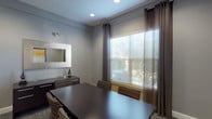 This compact yet well-appointed conference room offers a polished, professional environment designed for focused meetings and collaboration. The space is finished in a calming cool gray palette that lends a modern, neutral backdrop to all interior elements. At the center of the room sits a substantial dark espresso-toned rectangular conference table surrounded by upholstered chairs featuring a distinctive geometric patterned fabric in earth tones, providing both comfort and visual interest.

Natural light filters generously into the room through a large window dressed with layered window treatments — horizontal blinds for light control paired with floor-length dark charcoal curtains that frame the view of surrounding greenery. Recessed ceiling lighting supplements the natural light, ensuring consistent illumination throughout the day.

Along one wall, a dark credenza provides functional storage and display space, accented by decorative sculptural pieces. A large framed mirror with a metallic border is mounted above, adding depth and dimension to the room. On the opposite wall, a bold abstract artwork featuring warm gold and white tones serves as a striking focal point, adding an artistic and contemporary touch.

The flooring is finished with a patterned commercial-grade carpet in warm neutral tones, grounding the space practically and aesthetically. A private entrance door completes this self-contained, fully enclosed meeting room.