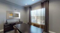 This compact yet well-appointed conference room offers a polished, professional environment designed for focused meetings and collaboration. The space is finished in a calming cool gray palette that lends a modern, neutral backdrop to all interior elements. At the center of the room sits a substantial dark espresso-toned rectangular conference table surrounded by upholstered chairs featuring a distinctive geometric patterned fabric in earth tones, providing both comfort and visual interest.

Natural light filters generously into the room through a large window dressed with layered window treatments — horizontal blinds for light control paired with floor-length dark charcoal curtains that frame the view of surrounding greenery. Recessed ceiling lighting supplements the natural light, ensuring consistent illumination throughout the day.

Along one wall, a dark credenza provides functional storage and display space, accented by decorative sculptural pieces. A large framed mirror with a metallic border is mounted above, adding depth and dimension to the room. On the opposite wall, a bold abstract artwork featuring warm gold and white tones serves as a striking focal point, adding an artistic and contemporary touch.

The flooring is finished with a patterned commercial-grade carpet in warm neutral tones, grounding the space practically and aesthetically. A private entrance door completes this self-contained, fully enclosed meeting room.