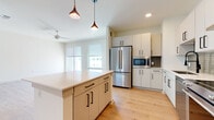 This contemporary single-floor residence showcases a thoughtfully designed open-concept layout with a clean, modern aesthetic throughout. The kitchen is a standout feature, equipped with stainless steel appliances including a French door refrigerator, built-in microwave, dishwasher, and a professional-grade range topped by a stainless steel hood. Flat-front cabinetry in a warm off-white tone is paired with matte black hardware, while a subway tile backsplash and a generous center island with a quartz countertop add both style and function. Pendant lighting with copper-toned fixtures illuminates the space, complementing the warm light wood flooring that flows throughout the main living areas. The living area is bright and airy, benefiting from large windows with roller shades and a ceiling fan, with access to a balcony beyond. Bedrooms feature plush carpet flooring and generous wall space. The primary bathroom is elegantly finished with large-format tile flooring, a glass-enclosed shower with marble-style tile, a dual-sink vanity with a white countertop and matte black fixtures, and a framed mirror. A walk-in closet is conveniently accessible directly from the bathroom. Overall, the space exudes a fresh, move-in-ready quality with consistent modern finishes and abundant natural light.