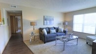 This single-floor residence offers a thoughtfully designed layout that balances comfort and style. The living area serves as the heart of the home, featuring plush wall-to-wall carpeting that adds warmth underfoot, complemented by smooth, neutral-toned walls that create a clean and calming backdrop. A textured ceiling adds subtle architectural character to the space. Natural light filters in through a window, brightening the room and enhancing its open feel. The living area is well-proportioned, providing ample space for relaxed seating and entertaining. The transition from hardwood-style flooring at the entryway to soft carpeting in the main living space clearly defines the functional zones within the home. Decorative touches, including tasteful wall art and coordinated accent lamps with metallic finishes, lend the space a polished, contemporary aesthetic. A glass-top coffee table and matching side table reflect light and contribute to an airy, modern sensibility. Overall, the space presents a cohesive and inviting atmosphere, combining practical design with refined decorative elements that make it both functional and visually appealing.