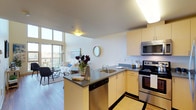 This single-floor residence is designed with an open-concept layout that seamlessly connects the kitchen and living areas, creating a cohesive and functional living environment. The kitchen is well-appointed with light wood cabinetry, dark countertops, a double-basin sink, a built-in dishwasher, and stainless steel appliances including a range and microwave. A peninsula counter extends the workspace and provides a natural division between the kitchen and the main living area, ideal for casual dining or meal prep. The living area is bathed in abundant natural light, courtesy of expansive floor-to-ceiling windows that frame views of the surrounding landscape. The light-toned hardwood flooring runs throughout, enhancing the sense of openness and warmth. Neutral walls and smooth ceilings contribute to a clean, modern aesthetic, while the generous window wall allows daylight to flood the interior throughout the day. The overall ambiance is bright, airy, and welcoming, with a contemporary design sensibility that balances style and practicality. The thoughtful layout and quality finishes make this a comfortable and visually appealing space suited for modern living.