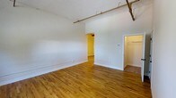 This single-floor space showcases a blend of industrial and classic design elements that create a warm, functional environment. The main room is defined by rich, honey-toned hardwood floors that run throughout, adding warmth and character to the open layout. Painted white brick walls lend an industrial charm while keeping the space feeling bright and airy. Overhead, exposed copper-toned pipes run horizontally across the upper portion of the room, adding a distinctive industrial accent consistent with loft-style architecture.

The elevated ceiling height enhances the sense of openness, making the main living area feel generously proportioned. To one side, an open doorway leads to an additional room bathed in warm yellow light, suggesting versatile use as a bedroom, office, or secondary living space. Adjacent to this doorway, a built-in closet with visible shelving and a rod provides practical storage. The clean white walls and trim throughout create a neutral backdrop that complements the natural materials, offering a move-in ready canvas adaptable to a variety of interior styles.