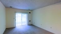 This single-floor residential space offers a clean, functional layout designed for comfortable everyday living. The main living area is finished with wall-to-wall carpeting that adds warmth underfoot, while smooth, neutral-toned walls provide a versatile backdrop for a variety of interior styles. A prominent sliding glass door fitted with vertical blinds serves as both a source of abundant natural light and a direct connection to the outdoors, offering pleasant views of the surrounding greenery. The textured popcorn ceiling adds a classic residential character to the space. Practical details include visible HVAC vents positioned both on the ceiling and lower on the walls, ensuring efficient climate control throughout. The room also features standard electrical outlets conveniently placed along the baseboards. The overall ambiance is bright, open, and move-in ready, making it a straightforward yet inviting space well-suited for a range of furnishing arrangements and personal touches.