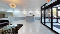 This single-floor commercial lobby space presents a sophisticated and welcoming environment, thoughtfully designed to balance functionality with contemporary style. The expansive main area is anchored by gleaming polished terrazzo floors that reflect the abundant recessed lighting overhead, creating a bright and open atmosphere throughout.

A standout feature is the curved, light grey reception desk with a white quartz countertop, positioned centrally to serve as both a functional workstation and a visual focal point. The desk's graceful form is complemented by matching grey beadboard wainscoting that lines the lower walls, adding texture and architectural character to the space.

Seating areas are thoughtfully arranged to accommodate visitors, featuring navy blue leather chairs and a curved velvet sofa, each paired with sleek gold-framed glass side tables. A bold black and gold starburst-patterned area rug grounds one of the seating vignettes, adding a dramatic design element.

Decorative gold sputnik chandeliers and warm wall sconces provide ambient lighting that enhances the space's upscale character. Large floor-to-ceiling glass doors and windows allow natural light to pour in, offering views to the exterior streetscape. Full-length mirrors along one wall visually expand the space, amplifying its sense of openness. The overall ambiance is polished, professional, and inviting.