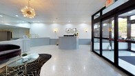 This single-floor commercial lobby space presents a sophisticated and welcoming environment, thoughtfully designed to balance functionality with contemporary style. The expansive main area is anchored by gleaming polished terrazzo floors that reflect the abundant recessed lighting overhead, creating a bright and open atmosphere throughout.

A standout feature is the curved, light grey reception desk with a white quartz countertop, positioned centrally to serve as both a functional workstation and a visual focal point. The desk's graceful form is complemented by matching grey beadboard wainscoting that lines the lower walls, adding texture and architectural character to the space.

Seating areas are thoughtfully arranged to accommodate visitors, featuring navy blue leather chairs and a curved velvet sofa, each paired with sleek gold-framed glass side tables. A bold black and gold starburst-patterned area rug grounds one of the seating vignettes, adding a dramatic design element.

Decorative gold sputnik chandeliers and warm wall sconces provide ambient lighting that enhances the space's upscale character. Large floor-to-ceiling glass doors and windows allow natural light to pour in, offering views to the exterior streetscape. Full-length mirrors along one wall visually expand the space, amplifying its sense of openness. The overall ambiance is polished, professional, and inviting.