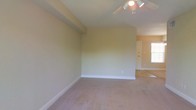 This single-floor space presents a clean, functional layout anchored by a spacious main room finished with wall-to-wall carpeting in a neutral tone. The room is fitted with a ceiling fan equipped with integrated lighting, providing both air circulation and overhead illumination. Smooth, light-colored walls are complemented by crisp white baseboards, lending the space a tidy, well-maintained appearance. A structural beam detail is visible along the upper wall, adding a subtle architectural element to the room. An open doorway leads to an adjoining area, where a white door and a window admitting natural daylight are visible, suggesting additional functional space beyond the main room. The overall ambiance is understated and versatile, offering a neutral canvas well-suited to a variety of furnishing arrangements and uses.