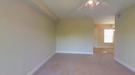 This single-floor space presents a clean, functional layout anchored by a spacious main room finished with wall-to-wall carpeting in a neutral tone. The room is fitted with a ceiling fan equipped with integrated lighting, providing both air circulation and overhead illumination. Smooth, light-colored walls are complemented by crisp white baseboards, lending the space a tidy, well-maintained appearance. A structural beam detail is visible along the upper wall, adding a subtle architectural element to the room. An open doorway leads to an adjoining area, where a white door and a window admitting natural daylight are visible, suggesting additional functional space beyond the main room. The overall ambiance is understated and versatile, offering a neutral canvas well-suited to a variety of furnishing arrangements and uses.
