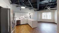 This single-floor unit showcases a striking industrial-modern aesthetic defined by its dramatic exposed ceiling, where black steel trusses, ductwork, and corrugated metal panels rise overhead, creating a loft-like atmosphere with impressive vertical volume. Rich wood-tone flooring runs throughout, providing a warm contrast to the crisp white walls and dark structural elements above.

The open-concept kitchen is a standout feature, outfitted with white shaker-style cabinetry, quartz countertops, and a bold white subway tile backsplash accented with black tile insets. A suite of stainless steel appliances — including a French door refrigerator, electric range, over-the-range microwave, and dishwasher — complement the modern design. A peninsula provides additional counter space and defines the transition into the living area.

The bedroom is generously proportioned, bathed in natural light through a large window with blinds, and topped with the same eye-catching exposed ceiling detail and a ceiling fan. A spacious walk-in closet with wire shelving and hanging rods offers substantial storage.

The bathroom continues the designer tilework theme, featuring a white-and-black tile surround, a white vanity with an integrated sink, and a glass-enclosed walk-in shower with dark tile walls. Every detail of this unit balances industrial character with contemporary comfort.
