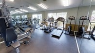 This fitness center offers a practical and well-organized workout environment designed to accommodate a range of exercise needs. The space is outfitted with a selection of cardio equipment, including treadmills and elliptical machines, positioned to take full advantage of the natural light streaming in through large windows that overlook surrounding greenery. The views of outdoor trees and landscaping create a pleasant backdrop during workouts.

Large floor-to-ceiling mirrors line one wall, visually expanding the space while allowing users to monitor their form. A weight bench and free weight equipment are positioned near the mirrored wall, providing a dedicated strength training area. The dark rubber flooring throughout is both durable and functional, offering cushioning and stability suited to high-impact exercise.

Overhead fluorescent lighting supplements the natural light, ensuring the space remains bright throughout the day. The walls feature a neutral two-tone color scheme with muted green and warm tan tones, contributing to a clean and focused atmosphere. The overall layout is efficient, allowing users to move freely between equipment without feeling cramped.