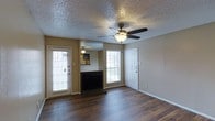 This 1-bedroom, 1-bathroom residence offers a thoughtfully organized single-floor layout that maximizes comfort and functionality. The living area serves as the heart of the home, showcasing rich hardwood-style flooring that extends throughout and lends a warm, inviting ambiance to the space. A ceiling fan with an integrated light fixture is centrally positioned overhead, providing both airflow and illumination beneath a textured ceiling. A fireplace with a dark surround serves as a natural focal point, flanked by large windows dressed with horizontal blinds that invite generous natural light into the room. A floor-to-ceiling mirror panel adjacent to the fireplace enhances the sense of openness and depth. Two exterior-facing doors provide convenient access and further contribute to the bright, airy feel of the living space. Neutral-toned walls throughout create a versatile backdrop well-suited to a variety of décor styles. The interior door leads seamlessly to the remaining areas of the home, maintaining a practical and connected flow. Overall, this residence combines classic design elements with functional features, resulting in a comfortable and well-lit living environment.