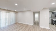 This single-floor unit offers three bedrooms, one bathroom, a living room, kitchen, and connecting hallway, all finished with a clean, contemporary aesthetic. Neutral warm-beige walls and white trim are consistent throughout, creating a cohesive and inviting atmosphere.

The living room is a comfortable open area featuring light wood-tone laminate flooring, recessed lighting, and a vertical-blind sliding door that allows natural light to filter in. A hallway extends from the living area, leading to the remaining rooms via white-paneled doors with matte black hardware.

The kitchen is designed as a galley-style space with dark gray shaker cabinets, white countertops, a stainless steel sink, subway tile backsplash, and stainless steel appliances including a refrigerator, range, and microwave.

The three bedrooms feature plush neutral carpet, windowed walls with blinds providing natural light, and clean wall finishes. The primary bedroom is a well-proportioned space offering comfortable room for furnishings.

The bathroom includes a tub-shower combination with a multi-toned linear tile surround, matte black fixtures, gray tile flooring, and crisp white walls, delivering a modern and polished finish.