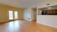 This single-floor space is designed with an open layout that promotes a spacious, airy feel throughout. The main living area is finished with warm-toned hardwood floors that run seamlessly across the space, contributing to a clean and cohesive aesthetic. Natural light flows generously through a set of glass-panel French doors along one wall, brightening the interior and providing a welcoming atmosphere.

The open floor plan allows for flexible furniture arrangements and easy movement throughout the space. A pass-through opening connects the living area to the kitchen, which features dark cabinetry with a contrasting dark countertop and a tile backsplash, visible from the main room. This design detail enhances the sense of connectivity between the living and kitchen areas while maintaining a degree of separation.

Additional features include a closeted storage area accessible from the main room, a wall-mounted thermostat, and standard HVAC vents integrated into the floor and ceiling. White trim and neutral wall tones complement the hardwood floors, creating a bright and versatile interior suitable for a variety of uses.