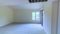 This single-story space is defined by its vaulted ceiling, which adds a sense of openness and volume to the interior. The ceiling features a prominent ventilation panel, contributing to effective climate control throughout the area. Neutral-toned walls and light beige wall-to-wall carpeting create a clean, understated palette that is versatile for a variety of uses and furnishing styles.

Natural light plays a significant role in the ambiance of this space. A large window on the far wall frames views of lush greenery outside, flooding the interior with bright, even daylight. Adjacent to the window, a sliding glass door with a sleek black frame provides both additional light and direct access to the outdoors, enhancing the connection between the interior and the surrounding environment.

The layout is open and unobstructed, offering generous usable floor area that can accommodate a wide range of configurations. White baseboards provide a clean finish along the perimeter, complementing the bright, airy feel of the space. Overall, the room presents a well-lit, functional environment with architectural details that elevate its character.
