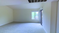 This single-story space is defined by its vaulted ceiling, which adds a sense of openness and volume to the interior. The ceiling features a prominent ventilation panel, contributing to effective climate control throughout the area. Neutral-toned walls and light beige wall-to-wall carpeting create a clean, understated palette that is versatile for a variety of uses and furnishing styles.

Natural light plays a significant role in the ambiance of this space. A large window on the far wall frames views of lush greenery outside, flooding the interior with bright, even daylight. Adjacent to the window, a sliding glass door with a sleek black frame provides both additional light and direct access to the outdoors, enhancing the connection between the interior and the surrounding environment.

The layout is open and unobstructed, offering generous usable floor area that can accommodate a wide range of configurations. White baseboards provide a clean finish along the perimeter, complementing the bright, airy feel of the space. Overall, the room presents a well-lit, functional environment with architectural details that elevate its character.