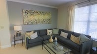 This single-floor space offers a thoughtfully designed living area that combines comfort and style in equal measure. The living room serves as the centerpiece, showcasing soft gray walls accented by crisp white crown molding that adds a refined architectural detail to the room. A large, sectional sofa in charcoal gray anchors the space, complemented by an assortment of coordinating throw pillows in muted greens, cream, and gold tones. A glass-top coffee table with a sleek metal frame sits at the center, adding a modern touch while maintaining an open, airy feel. Natural light filters through the windows, dressed with sheer cream drapery that softens the room's ambiance. A striking large-scale abstract painting in earthy yellows, greens, and blacks serves as a bold focal point above the sofa, infusing the space with artistic character. Decorative vases and accent pieces in complementary tones are thoughtfully arranged throughout. The neutral-toned flooring ties the room together, creating a cohesive and inviting atmosphere. Overall, the space presents a balanced blend of contemporary design elements and warm, livable comfort.